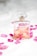 A clear glass perfume bottle with a light pink liquid inside is surrounded by scattered pink rose petals on a soft white surface. The bottle has a square shape with a gold label and a transparent top, conveying a sense of luxury and elegance.