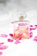 A clear glass perfume bottle with a light pink liquid inside is surrounded by scattered pink rose petals on a soft white surface. The bottle has a square shape with a gold label and a transparent top, conveying a sense of luxury and elegance.