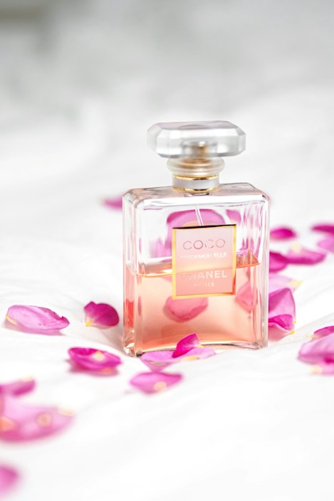 A clear glass perfume bottle with a light pink liquid inside is surrounded by scattered pink rose petals on a soft white surface. The bottle has a square shape with a gold label and a transparent top, conveying a sense of luxury and elegance.