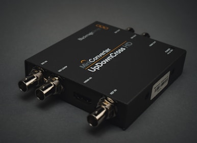 A black electronic device with various input and output ports labeled SDI IN, SDI LOOP, HDMI IN, and USB. The label on the device reads 'Mini Converter UpDownCross HD'. It is placed on a dark surface, and the connectors are visible in a metallic finish.