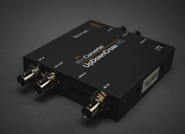 A black electronic device with various input and output ports labeled SDI IN, SDI LOOP, HDMI IN, and USB. The label on the device reads 'Mini Converter UpDownCross HD'. It is placed on a dark surface, and the connectors are visible in a metallic finish.