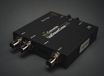 A black electronic device with various input and output ports labeled SDI IN, SDI LOOP, HDMI IN, and USB. The label on the device reads 'Mini Converter UpDownCross HD'. It is placed on a dark surface, and the connectors are visible in a metallic finish.