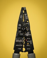 A pair of black wire strippers or crimpers with silver-gray handles against a solid yellow background. The tool displays various measurement markings and instructions for wire stripping, cutting, and crimping.