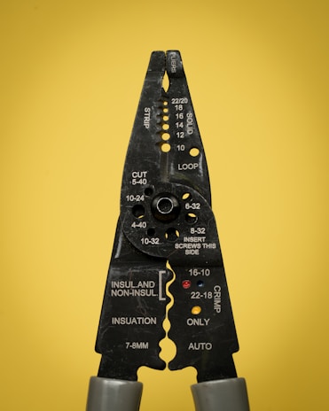 A pair of black wire strippers or crimpers with silver-gray handles against a solid yellow background. The tool displays various measurement markings and instructions for wire stripping, cutting, and crimping.