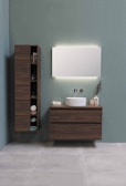 A modern bathroom setup with a minimalist aesthetic. A large wooden cabinet with open shelves holds various items, including neatly folded towels and toiletries. Adjacent, a floating wooden vanity with a round white basin sits beneath a large rectangular mirror that is softly backlit. The background is a calm, muted green wall, and the floor consists of large, light gray tiles.