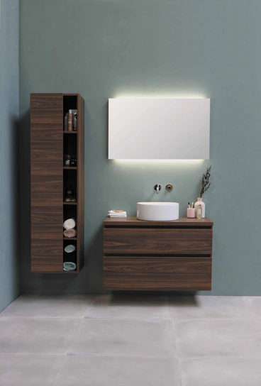 A modern bathroom setup with a minimalist aesthetic. A large wooden cabinet with open shelves holds various items, including neatly folded towels and toiletries. Adjacent, a floating wooden vanity with a round white basin sits beneath a large rectangular mirror that is softly backlit. The background is a calm, muted green wall, and the floor consists of large, light gray tiles.