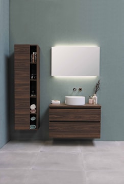 A modern bathroom setup with a minimalist aesthetic. A large wooden cabinet with open shelves holds various items, including neatly folded towels and toiletries. Adjacent, a floating wooden vanity with a round white basin sits beneath a large rectangular mirror that is softly backlit. The background is a calm, muted green wall, and the floor consists of large, light gray tiles.