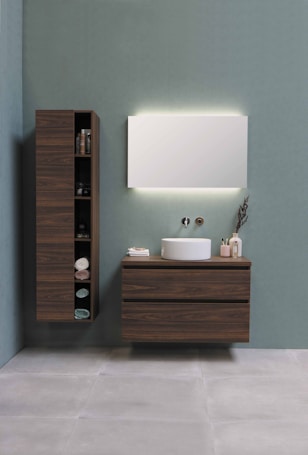 A modern bathroom setup with a minimalist aesthetic. A large wooden cabinet with open shelves holds various items, including neatly folded towels and toiletries. Adjacent, a floating wooden vanity with a round white basin sits beneath a large rectangular mirror that is softly backlit. The background is a calm, muted green wall, and the floor consists of large, light gray tiles.