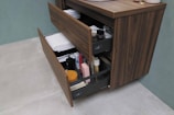 A wooden cabinet with open drawers reveals neatly arranged towels, various toiletries, and personal care items. The drawers contain a mix of folded white towels, bottles, a sponge, and boxes. The cabinet is placed against a green wall and stands on a light gray tiled floor.