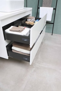 Two open drawers with neatly arranged towels and toiletries in a modern bathroom setting. A white countertop with a sink is present above the drawers. In the background, there is a hanging towel on a black metal rack.
