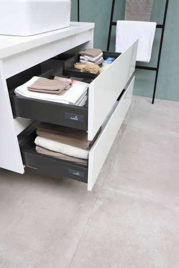 Two open drawers with neatly arranged towels and toiletries in a modern bathroom setting. A white countertop with a sink is present above the drawers. In the background, there is a hanging towel on a black metal rack.