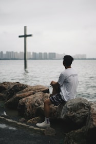 man in white shirt sitting on brown rock near body of water during daytime