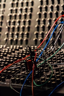 A close-up view of a sound or patch panel filled with numerous small connectors or jacks, intertwined with several colored wires including red, blue, black, and green. The background is filled with multiple similar connectors, creating a dense and complex texture.