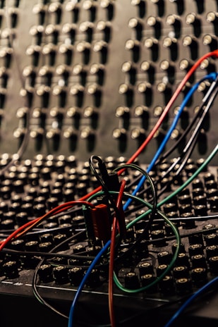 A close-up view of a sound or patch panel filled with numerous small connectors or jacks, intertwined with several colored wires including red, blue, black, and green. The background is filled with multiple similar connectors, creating a dense and complex texture.