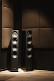 Two tall black speakers placed against a wall with dark brown acoustic panels. Wooden flooring provides a warm contrast to the otherwise dimly lit room.