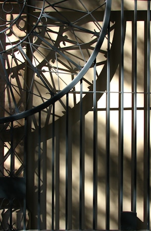 Intricate metalwork casts complex geometric shadows on a sunlit beige wall, creating an interplay of light and shadow with diagonal and circular patterns.