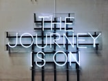 A neon sign displays the words 'THE JOURNEY IS ON' in bright white letters against a grid-like background mounted on a plain wall. The design features a modern, industrial aesthetic with a minimalist approach.