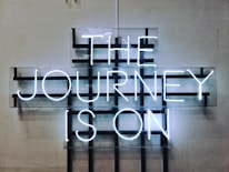 A neon sign displays the words 'THE JOURNEY IS ON' in bright white letters against a grid-like background mounted on a plain wall. The design features a modern, industrial aesthetic with a minimalist approach.
