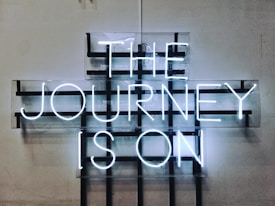 A neon sign displays the words 'THE JOURNEY IS ON' in bright white letters against a grid-like background mounted on a plain wall. The design features a modern, industrial aesthetic with a minimalist approach.
