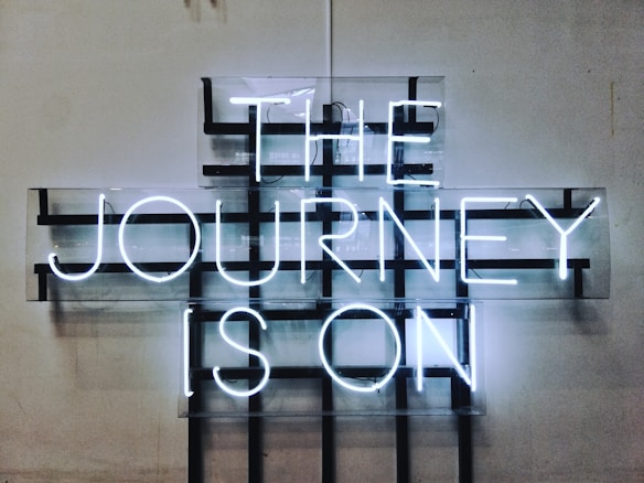 A neon sign displays the words 'THE JOURNEY IS ON' in bright white letters against a grid-like background mounted on a plain wall. The design features a modern, industrial aesthetic with a minimalist approach.