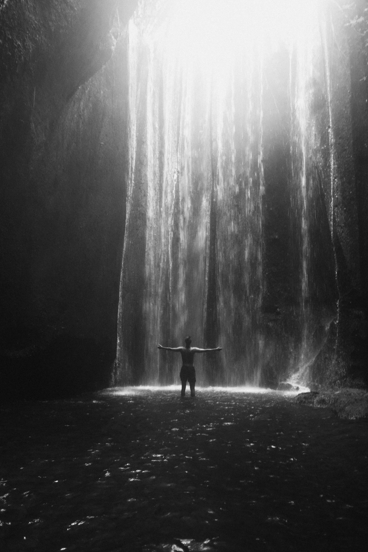 A person standing under the Tukad Cepung waterfall with arms out, light falling through the cave roof