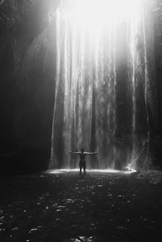 A person stands with arms outstretched under a cascading waterfall, surrounded by the dark, rocky walls of a cave under a bright light shining from above.
