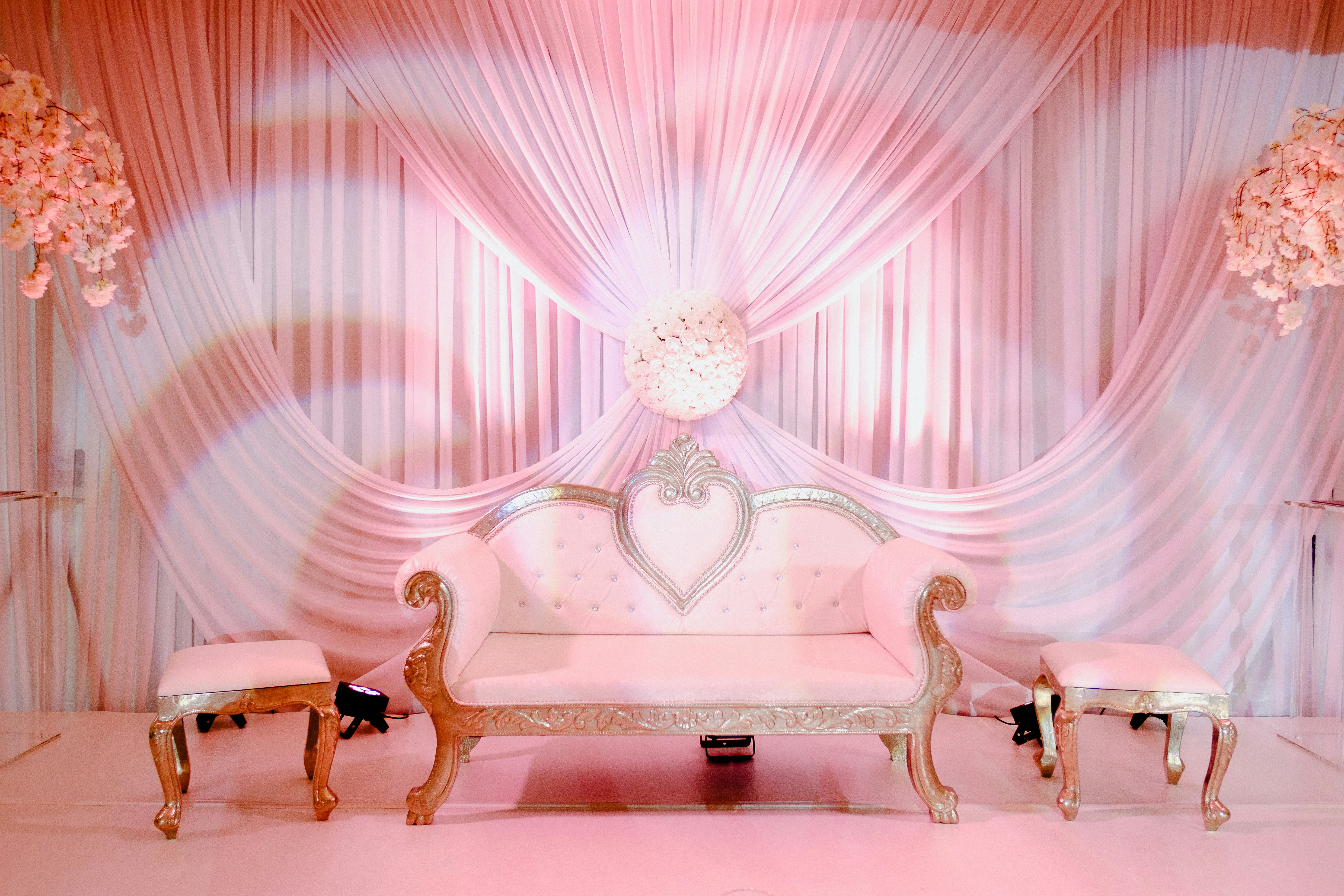 A luxurious white sofa with ornate gold detailing is positioned centrally, flanked by two matching stools. The backdrop consists of elegant, draped fabric in soft pink hues, complemented by floral arrangements on either side.
