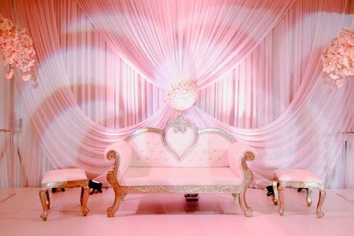 A luxurious white sofa with ornate gold detailing is positioned centrally, flanked by two matching stools. The backdrop consists of elegant, draped fabric in soft pink hues, complemented by floral arrangements on either side.