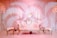 A luxurious white sofa with ornate gold detailing is positioned centrally, flanked by two matching stools. The backdrop consists of elegant, draped fabric in soft pink hues, complemented by floral arrangements on either side.