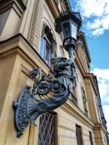 An ornate black metal sculpture adorns the fa&ccedil;ade of a classic architectural building. The sculpture portrays an intricate design featuring a figure holding an elaborate lantern, its detail contrasting with the light brown exterior walls adorned with decorative molding around windows.