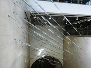 An architectural installation features numerous thin, elongated metallic rods arranged horizontally and diagonally along a wall in a large, open interior space. The ceiling is adorned with hanging lights and there is an escalator visible at the lower portion of the image. The setting appears modern and industrial.