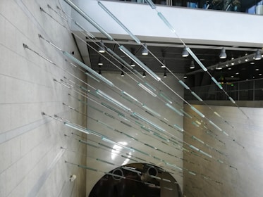 An architectural installation features numerous thin, elongated metallic rods arranged horizontally and diagonally along a wall in a large, open interior space. The ceiling is adorned with hanging lights and there is an escalator visible at the lower portion of the image. The setting appears modern and industrial.
