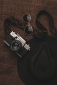 A vintage camera with a woven strap is placed on a textured brown surface alongside a pair of aviator sunglasses and a wide-brimmed hat.