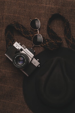 A vintage camera with a woven strap is placed on a textured brown surface alongside a pair of aviator sunglasses and a wide-brimmed hat.