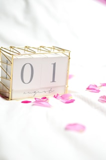 A decorative desk calendar displaying the date '01 August' in a gold wireframe holder. The calendar is placed on a white surface scattered with pink rose petals, creating a delicate and romantic atmosphere.