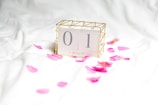 A minimalist calendar block displaying the date April 1, surrounded by scattered pink rose petals on a white fabric background. The calendar is encased in a gold wire frame, adding a touch of elegance.