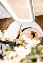 A woman is reaching for a baguette in a modern bakery setting, surrounded by various baked goods and a stylishly lit interior. The bakery display case is filled with bread and other items wrapped in clear packaging.