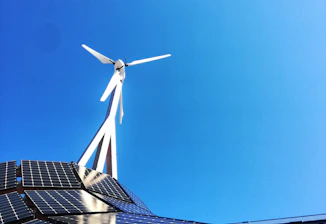 A clear blue sky provides the backdrop for a modern renewable energy setup featuring a wind turbine and solar panels. The sleek, geometric arrangement of solar panels reflects light while the blades of the wind turbine cut through the sky above.