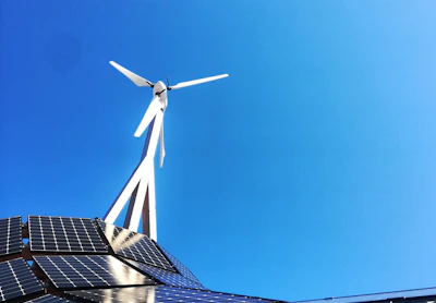 A clear blue sky provides the backdrop for a modern renewable energy setup featuring a wind turbine and solar panels. The sleek, geometric arrangement of solar panels reflects light while the blades of the wind turbine cut through the sky above.
