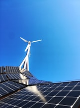 A clear blue sky provides the backdrop for a modern renewable energy setup featuring a wind turbine and solar panels. The sleek, geometric arrangement of solar panels reflects light while the blades of the wind turbine cut through the sky above.