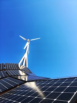 A clear blue sky provides the backdrop for a modern renewable energy setup featuring a wind turbine and solar panels. The sleek, geometric arrangement of solar panels reflects light while the blades of the wind turbine cut through the sky above.