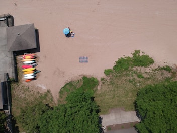 An aerial view of a sandy beach with a small arrangement of kayaks in various colors near a building. A blue umbrella provides shade for two people sitting on beach chairs. A striped blanket lies on the sand, and there are patches of greenery along the edge of the beach.