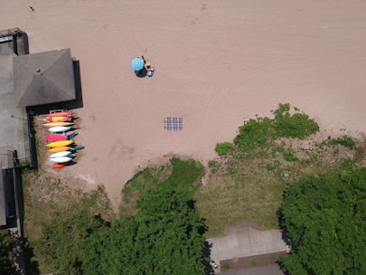 An aerial view of a sandy beach with a small arrangement of kayaks in various colors near a building. A blue umbrella provides shade for two people sitting on beach chairs. A striped blanket lies on the sand, and there are patches of greenery along the edge of the beach.