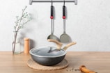 On a wooden surface, a glass vase with a flowering branch stands beside a pair of wooden salt and pepper shakers. In the foreground, there's a lidded frying pan with wooden handles resting on a braided mat. Two kitchen utensils hang on a wall-mounted rack above.