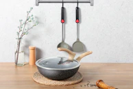 On a wooden surface, a glass vase with a flowering branch stands beside a pair of wooden salt and pepper shakers. In the foreground, there's a lidded frying pan with wooden handles resting on a braided mat. Two kitchen utensils hang on a wall-mounted rack above.