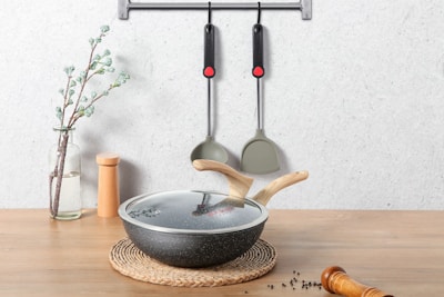 On a wooden surface, a glass vase with a flowering branch stands beside a pair of wooden salt and pepper shakers. In the foreground, there's a lidded frying pan with wooden handles resting on a braided mat. Two kitchen utensils hang on a wall-mounted rack above.