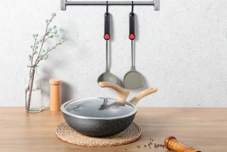 On a wooden surface, a glass vase with a flowering branch stands beside a pair of wooden salt and pepper shakers. In the foreground, there's a lidded frying pan with wooden handles resting on a braided mat. Two kitchen utensils hang on a wall-mounted rack above.