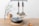 On a wooden surface, a glass vase with a flowering branch stands beside a pair of wooden salt and pepper shakers. In the foreground, there's a lidded frying pan with wooden handles resting on a braided mat. Two kitchen utensils hang on a wall-mounted rack above.