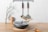 On a wooden surface, a glass vase with a flowering branch stands beside a pair of wooden salt and pepper shakers. In the foreground, there's a lidded frying pan with wooden handles resting on a braided mat. Two kitchen utensils hang on a wall-mounted rack above.