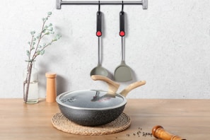 On a wooden surface, a glass vase with a flowering branch stands beside a pair of wooden salt and pepper shakers. In the foreground, there's a lidded frying pan with wooden handles resting on a braided mat. Two kitchen utensils hang on a wall-mounted rack above.